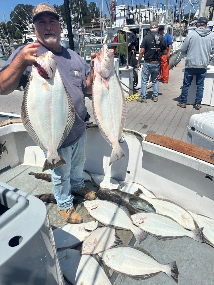 Man holding two large fish on a boat with more fish at his feet, other people in the background.